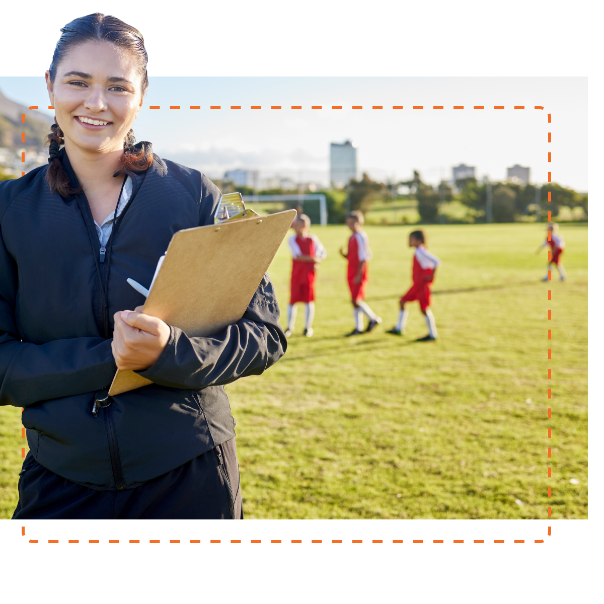 A female soccer coach smiling at the camera, clipboard in hand, with young players in red uniforms practicing on a sunny field in the background, suggesting a well-organized team environment facilitated by engaged coaching.